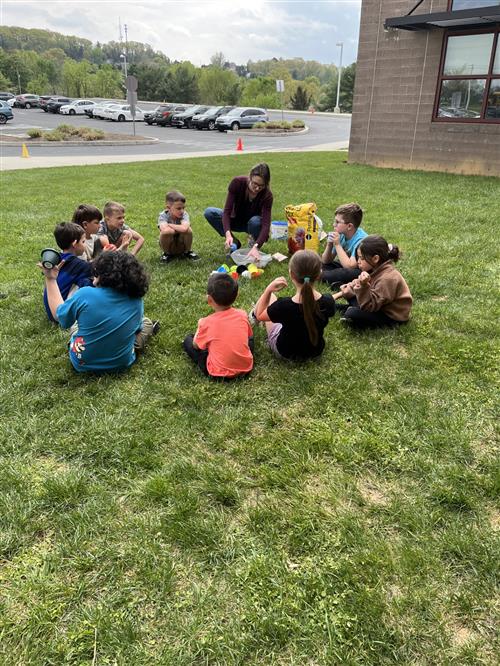 Students sitting in grass with teacher holding flower pots